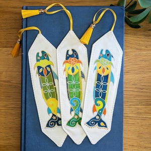 May include: Three white fabric bookmarks, each featuring a colorful, stylized fish illustration in shades of blue, green, yellow, and orange. Each bookmark is topped with a bright yellow tassel attached by a thin cord, creating a decorative set.