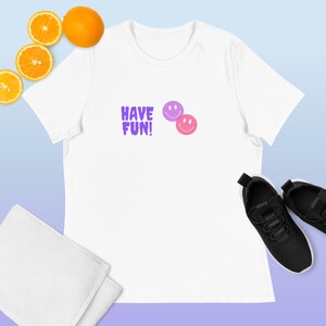 Puede incluir: Camiseta blanca con el texto "HAVE FUN!" en morado, junto con dos emoticonos sonrientes, uno rosa y otro morado. La imagen incluye zapatillas negras y rodajas de naranja.