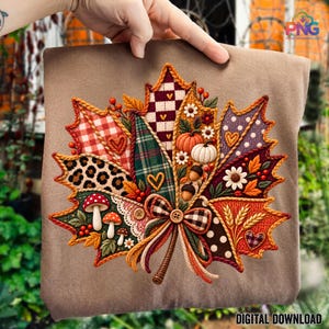 May include: A tan t-shirt featuring a large, detailed embroidered-style graphic of a maple leaf. The leaf is divided into sections with various patterns including red plaid, leopard print, polka dots, and checkered designs, accented with mushrooms, pumpkins, acorns, and a central bow.