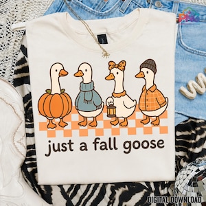 Puede incluir: Una camiseta color crema con un gráfico de cuatro gansos sobre un patrón de cuadros naranjas y blancos. Los gansos llevan ropa de temática otoñal, como un disfraz de calabaza, una bufanda azul, un lazo y una camisa de cuadros. El texto dice: just a fall goose.