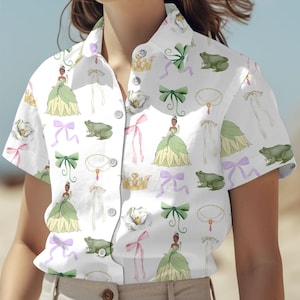 May include: A white button-down shirt with a short sleeve featuring a repeating pattern of a cartoon princess, frogs, flowers, crowns, and bows. The princess is wearing a green dress and a crown.