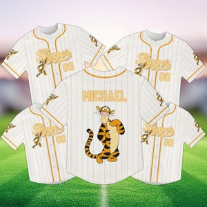 Könnte beinhalten: Weiße Baseball-Trikots mit orangefarbenen Nadelstreifen und dem Namen "Tigger" in goldener Schrift. Ein Trikot hat "MICHAEL" auf dem Rücken und eine Tigger-Cartoon-Grafik. Die Trikots haben die Nummer "00".