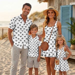 May include: A family wearing matching white clothing with a black Mickey Mouse head pattern. The adult wears a button-down shirt and light trousers, the child wears a button-down shirt and shorts, and the two others wear sleeveless dresses.