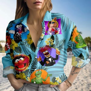 Peut inclure: Chemise bleu clair à boutons avec un imprimé vibrant de personnages des Muppets, dont Miss Piggy et Kermit la Grenouille. La chemise présente un motif floral tropical avec des accents jaunes et verts.