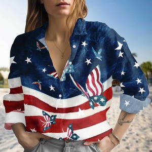 May include: Button-down shirt with a patriotic design featuring the American flag and butterflies. The shirt has a blue background with white stars and red and white stripes. The butterflies have a similar patriotic design.