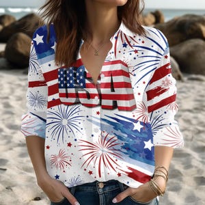 May include: White button-down shirt with a patriotic USA theme. The shirt features the American flag, fireworks, and the word "USA" in bold letters. Red, white, and blue colors dominate the design, creating a festive look.