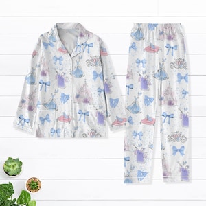 May include: A white pyjama set with a long-sleeved top and trousers. The pyjamas feature a repeating pattern of Cinderella, her castle, and blue bows.