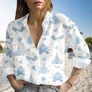 May include: White button-down shirt featuring a repeating pattern of blue Cinderella-themed illustrations, including castles, carriages, gowns, and glass slippers, with yellow star accents. The long-sleeved shirt is made of a lightweight fabric.