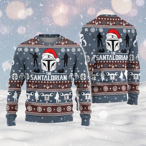 May include: A blue crew neck jumper with a festive design. The jumper features a Mandalorian helmet wearing a Santa hat, silhouettes, and the text "The Santalorian". The design includes snowflakes and brown accents.