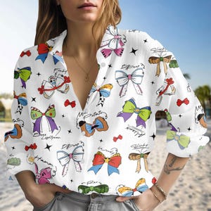 May include: White button-down shirt with a pattern of colourful bow illustrations and character names. The long-sleeved shirt has a classic collar and is made of a lightweight fabric. The design includes bows in various colours and styles.
