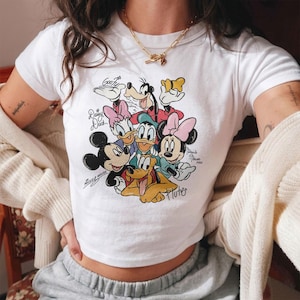 May include: White crop top featuring a colorful graphic of Disney characters including Mickey Mouse, Minnie Mouse, Donald Duck, Daisy Duck, Goofy, and Pluto. The shirt has short sleeves and a crew neck. The text "Mickey Mouse" and "Minnie Mouse" are also printed on the shirt.