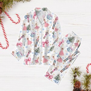 May include: White pajamas with a long-sleeved top and pants. The pajamas feature a repeating pattern of ballerinas, nutcrackers, and Christmas trees. The pattern is in shades of pink, blue, and gold.