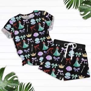 May include: A black and white short-sleeved shirt and matching shorts with a repeating pattern of a cartoon mermaid, seashells, crowns, and bows.