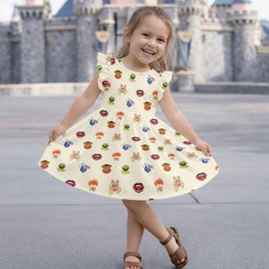 Puede incluir: Un vestido color crema con un estampado de caras de personajes de los Muppets. El vestido tiene mangas cortas con volantes y una falda acampanada. La niña lleva sandalias marrones.