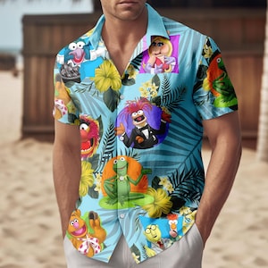 Op de afbeelding: Lichtblauw Hawaiiaans overhemd met verschillende Muppet-personages, gele bloemen en tropische bladeren. Het overhemd heeft een knoopsluiting en korte mouwen. De achtergrond is lichtblauw.