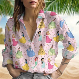 May include: A pink long-sleeved button-up shirt with a print of Disney princesses in various poses. The princesses are wearing colorful dresses and have blonde or brunette hair.