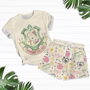 Puede incluir: Conjunto de camiseta y pantalón corto color crema. La camiseta presenta un escudo con un arpa, un libro, una cruz y un balón de fútbol. Los pantalones cortos tienen un patrón repetido con texto, coronas, corazones y el número 13. El texto incluye "Tommen College".