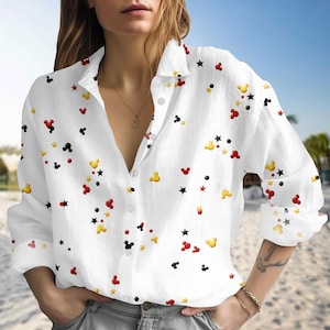 May include: White button-down shirt with a pattern of black, red, and yellow Mickey Mouse shapes and stars. The long-sleeved shirt has a classic collar and is made of a lightweight fabric. The shirt is unbuttoned at the top.
