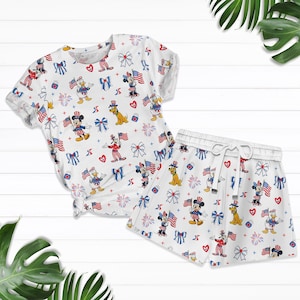 Op de afbeelding: Witte pyjamaset met een herhaald patroon van stripfiguren, Amerikaanse vlaggen, strikken en vuurwerk in rood, wit en blauw. De set bevat een top met korte mouwen en bijpassende shorts met een trekkoord in de taille.