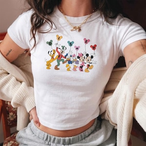 May include: White crop top featuring a colorful graphic of Disney characters, including Mickey and Minnie Mouse, holding balloons that spell out "DISNEY". The design is in a cartoon style with bright colors.