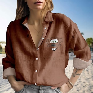 Puede incluir: Camisa marrón con botones y un dibujo de un coche de dibujos animados en el pecho. La camisa tiene mangas largas con puños doblados. La camisa está hecha de una tela suave y texturizada y tiene botones blancos.