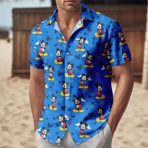 May include: Blue button-down shirt featuring a repeating pattern of Mickey Mouse characters in various poses. The shirt has short sleeves and a classic collar. Mickey Mouse is depicted in his iconic red shorts, yellow shoes, and white gloves. The background includes white hearts and Mickey Mouse silhouettes.