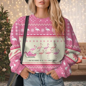 Puede incluir: Jersey navideño rosa con diseño de flamenco. El jersey presenta un fondo rosa con patrones de copos de nieve y flamencos blancos. El diseño central muestra un trineo tirado por flamencos, con el texto "I AM DREAMING OF A PINK CHRISTMAS!"