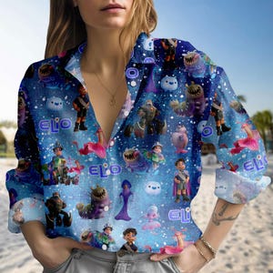 Może przedstawiać: Niebieska koszula zapinana na guziki z powtarzającym się wzorem postaci z kreskówek i napisem "Elio". Koszula ma długie rękawy i kołnierzyk. Tło jest jasnoniebieskie z białymi kropkami.