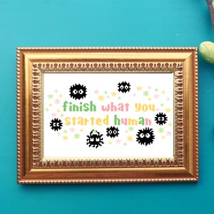 May include: Framed cross-stitch artwork featuring a white background with black and white pixelated characters and the text "finish what you started human". The characters are surrounded by colorful stars.