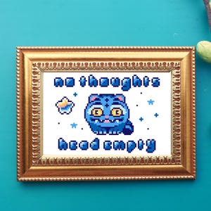 Può includere: Un'opera d'arte a punto croce incorniciata in una cornice dorata ornata. L'opera presenta un gatto pixelato blu con il testo "no thoughts head empty". Sono incluse anche stelle pixelate e un disegno a forma di stella.