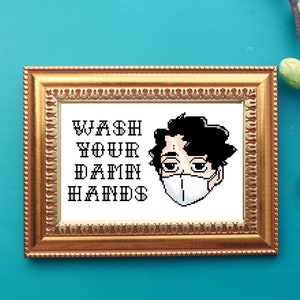 Puede incluir: Una obra de arte en punto de cruz enmarcada con un marco dorado ornamentado. La obra presenta el texto "WASH YOUR DAMN HANDS" sobre una ilustración pixelada de una persona con mascarilla. El fondo es de color turquesa vibrante.