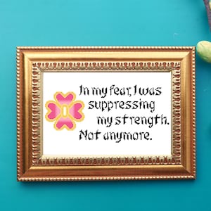 May include: A framed cross-stitch with a pink and yellow flower and the text "In my fear, I was suppressing my strength. Not anymore."