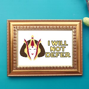 May include: A framed cross-stitch artwork with a gold-colored ornate frame. The artwork features a pixelated image of a face with a crown and the text "I WILL NOT DEFER" in yellow and black.