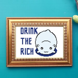 May include: A framed cross-stitch design with a white pixelated cartoon character and the text "Drink the Rich" in blue.