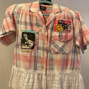 May include: A short-sleeved button-down shirt in a pink, white, and blue plaid pattern. It features a black and white photo patch labeled Donut Queen, a heart-shaped patch, a colorful button-adorned pocket patch, and a white lace-trimmed hem.