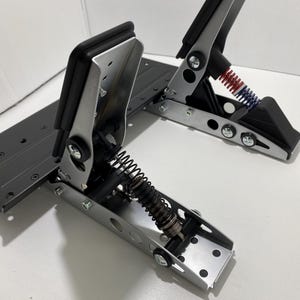 Peut inclure: Deux pédales de course argentées avec des coussinets noirs et des ressorts. Les pédales sont montées sur une base gris foncé. Une pédale a un ressort rouge et bleu, l'autre un ressort noir. Les pédales sont conçues pour le sim racing.