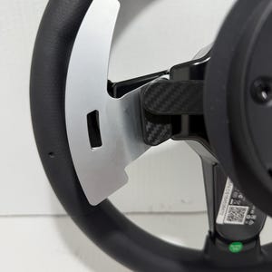 May include: Close-up of a black steering wheel with silver paddle shifters. The wheel has a textured grip and a carbon fibre accent. The paddle shifters are metallic and have a rectangular cutout.
