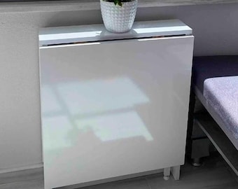 Puede incluir: Mesa plegable blanca con una maceta de cerámica blanca con una pequeña planta en la parte superior. La mesa está en una habitación con una ventana y un suelo de baldosas.