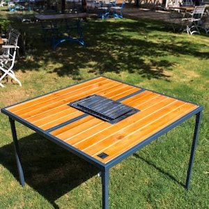 May include: Square outdoor table with a wooden top and a built-in grill. The tabletop is made of light brown wooden planks with a dark gray metal frame and legs. The table is set on a grassy lawn.