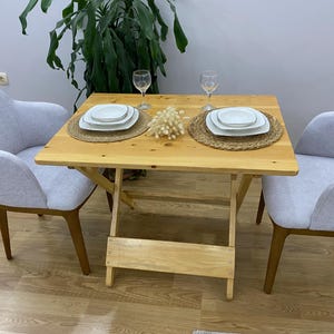 May include: A wooden folding table with two white chairs. The table is set with two place settings, each with a white plate, a white saucer, and a glass. The table is decorated with a small bouquet of dried flowers.