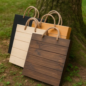Puede incluir: Cuatro bolsas de mano de madera en tonos negro, madera clara, madera media y madera oscura. Cada bolsa tiene forma rectangular con listones de madera horizontales y asas de cuerda. Las bolsas están apoyadas contra un tronco de árbol sobre una superficie cubierta de hierba.