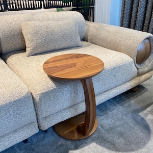 Puede incluir: Una mesa auxiliar de madera con una tapa redonda y una pata curva en forma de C. La mesa es de madera de tonos c&aacute;lidos y est&aacute; colocada junto a un sof&aacute; de color claro. El sof&aacute; tiene una tela texturizada y una almohada a juego.
