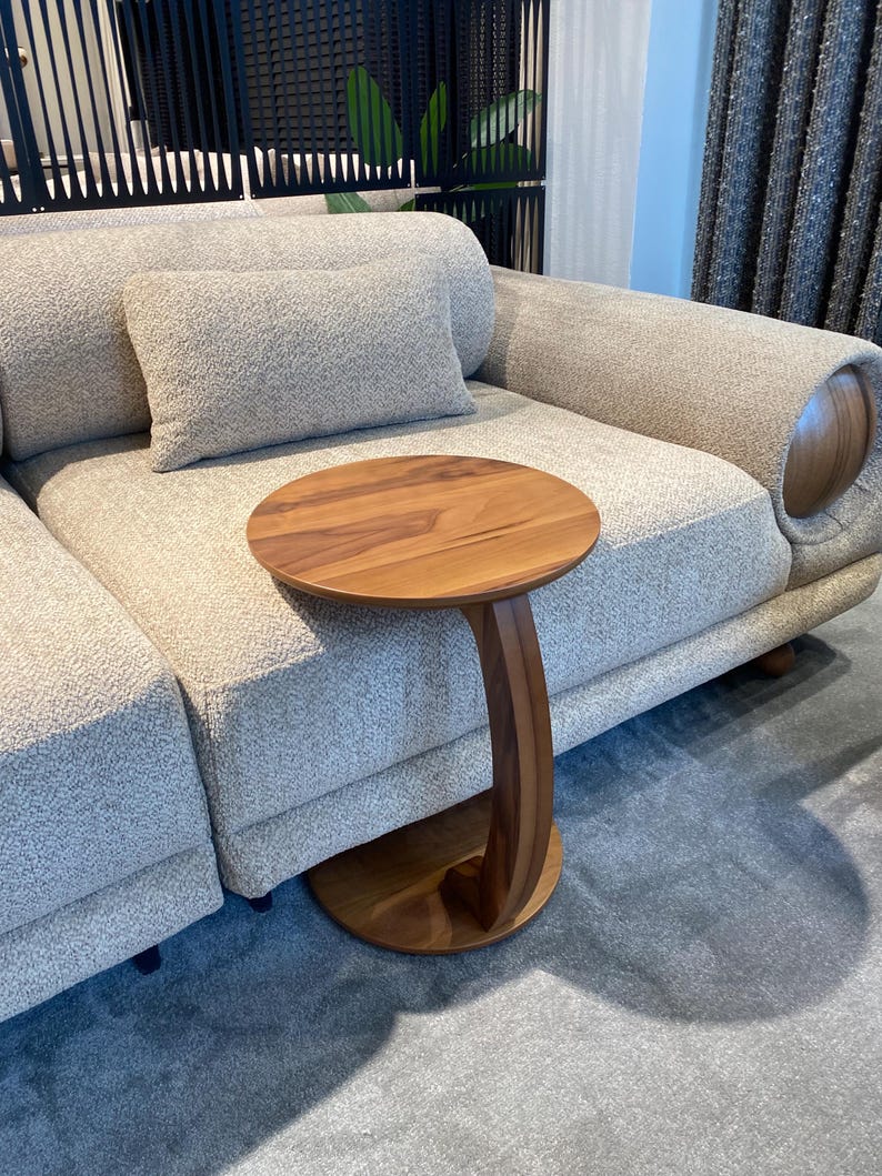 Puede incluir: Una mesa auxiliar de madera con una tapa redonda y una pata curva en forma de C. La mesa es de madera de tonos c&aacute;lidos y est&aacute; colocada junto a un sof&aacute; de color claro. El sof&aacute; tiene una tela texturizada y una almohada a juego.