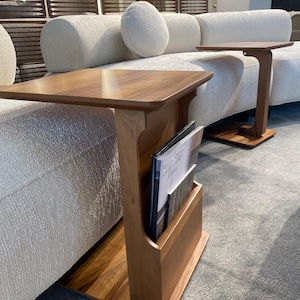 Puede incluir: Mesa auxiliar de madera con revistero. La mesa tiene una parte superior rectangular y un revistero vertical. La madera tiene un acabado c&aacute;lido y natural. La mesa est&aacute; colocada junto a un sof&aacute; de color claro.