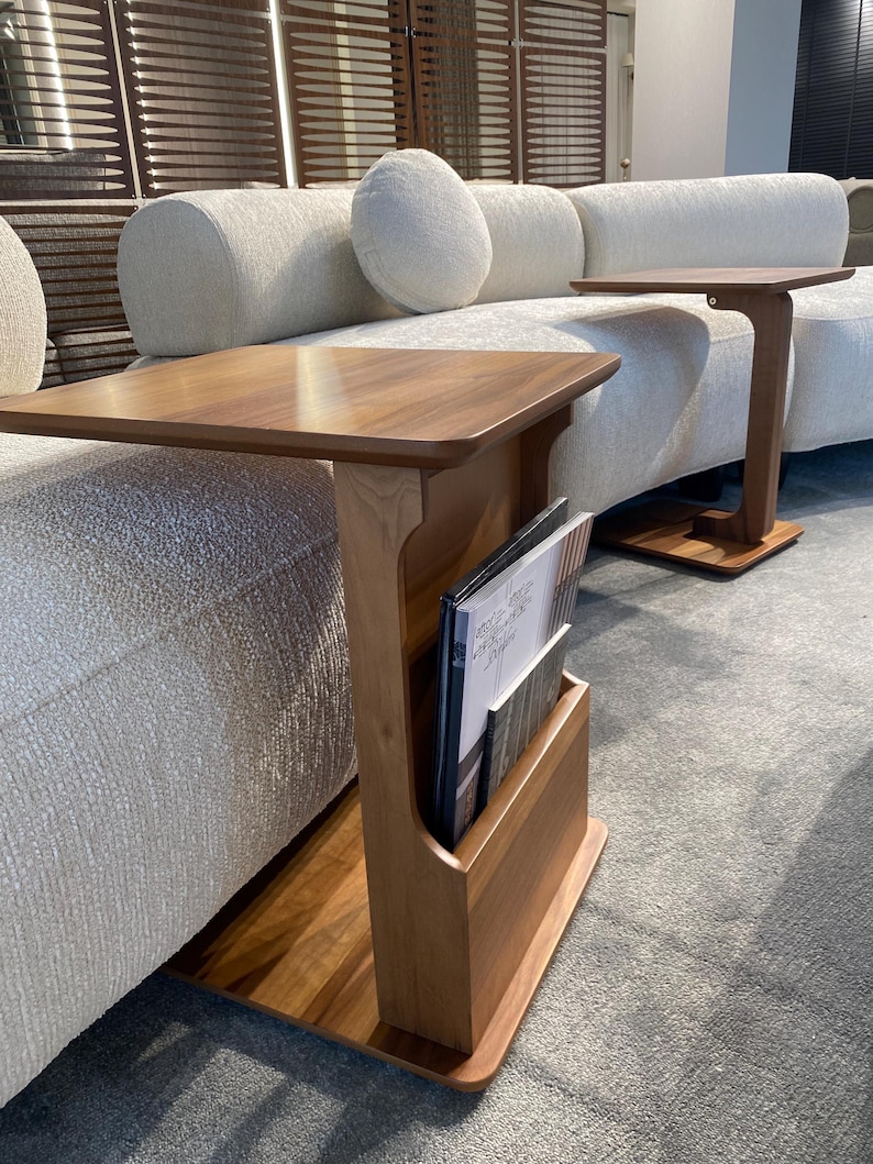 Puede incluir: Mesa auxiliar de madera con revistero. La mesa tiene una parte superior rectangular y un revistero vertical. La madera tiene un acabado c&aacute;lido y natural. La mesa est&aacute; colocada junto a un sof&aacute; de color claro.