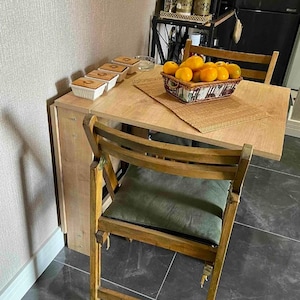 May include: A wooden folding table with a light wood finish and a green cushioned chair. The table is set with a basket of oranges and small white bowls.