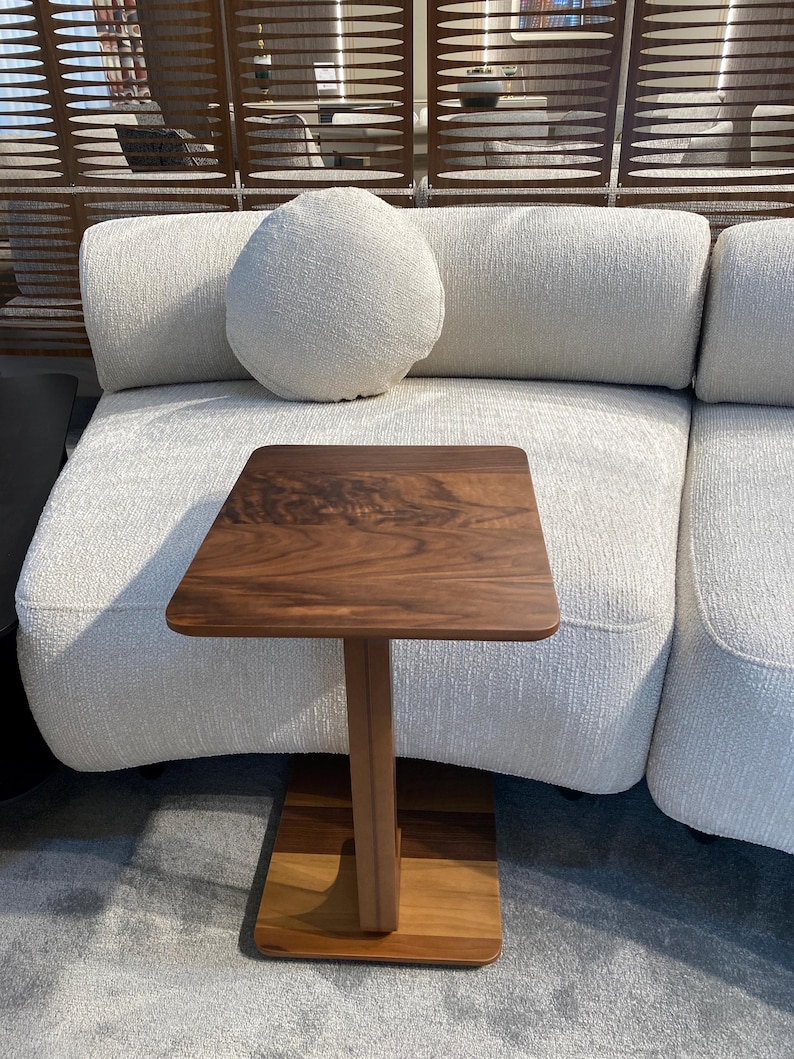Puede incluir: Una peque&ntilde;a mesa auxiliar cuadrada de madera con una tapa marr&oacute;n oscuro y una base marr&oacute;n m&aacute;s clara. La mesa tiene una sola pata y est&aacute; colocada frente a un sof&aacute; blanco texturizado con un coj&iacute;n redondo.