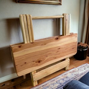 Puede incluir: Una mesa plegable de madera con un acabado de madera natural. La mesa está cerrada y plegada, con las patas metidas. La mesa está hecha de madera maciza y tiene un diseño simple y rústico.