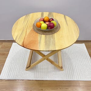 Pode incluir: Uma mesa redonda de madeira com acabamento em madeira natural. A mesa tem um design simples e rústico e é sustentada por uma base em forma de X robusta. Uma tigela de frutas fica sobre a mesa, adicionando um toque de cor e frescura.