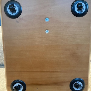 Puede incluir: Base de caja de madera marr&oacute;n claro con cuatro ruedas negras, una en cada esquina. Las ruedas tienen detalles blancos y negros. Dos peque&ntilde;os tornillos azul claro son visibles en el centro de la base.